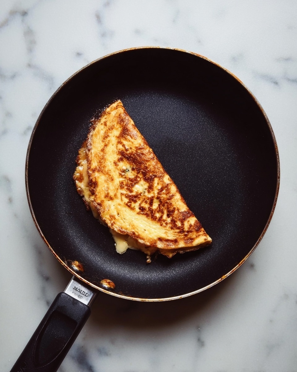 A black frying pan with a single folded golden-brown omelette in the center, showing a slightly crispy texture on the outside and some melted cheese peeking out from the edges, placed on a white marbled surface. photo taken with an iphone --ar 4:5 --v 7