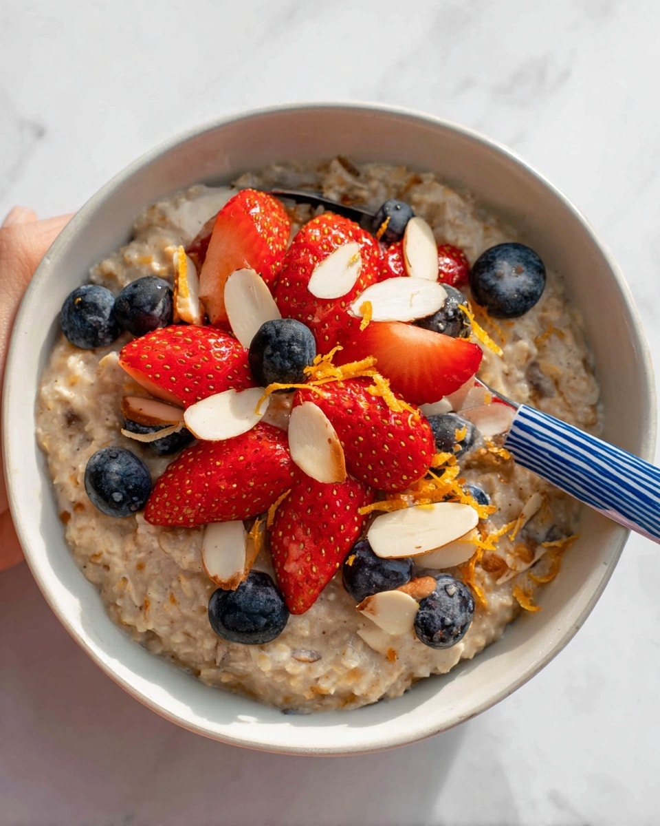 A close-up view of a bowl with three main layers: the bottom layer is creamy oatmeal with a light brown color and thick texture filling the bowl. On top of the oatmeal is a mix of fresh fruit, including bright red sliced strawberries and whole dark blue blueberries, arranged in the center. Thin, pale yellow almond slices are spread over the fruit, along with small sprinkled pieces of orange zest adding a bright touch. The bowl is white and sits on a white marbled surface. A woman’s hand is holding a spoon with blue and white stripes, placed inside the bowl. Photo taken with an iphone --ar 4:5 --v 7