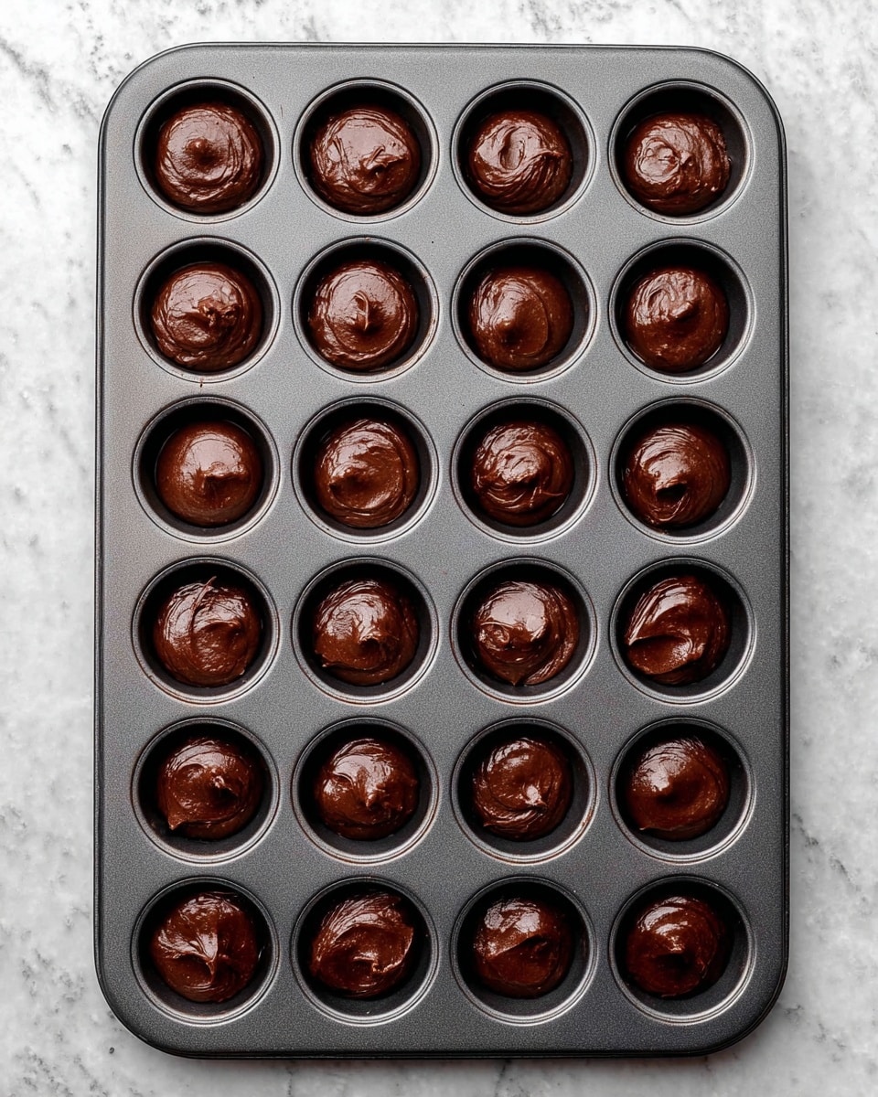 A dark gray baking tray with 24 round cups, each filled with smooth dark brown chocolate batter. The batter is slightly mounded above the cup edges with a glossy, thick texture that suggests it is ready to be baked. The tray rests on a white marbled surface, giving a clean and bright look. There are no other items or colors in the image, focusing only on the raw chocolate batter in the tray. photo taken with an iphone --ar 4:5 --v 7