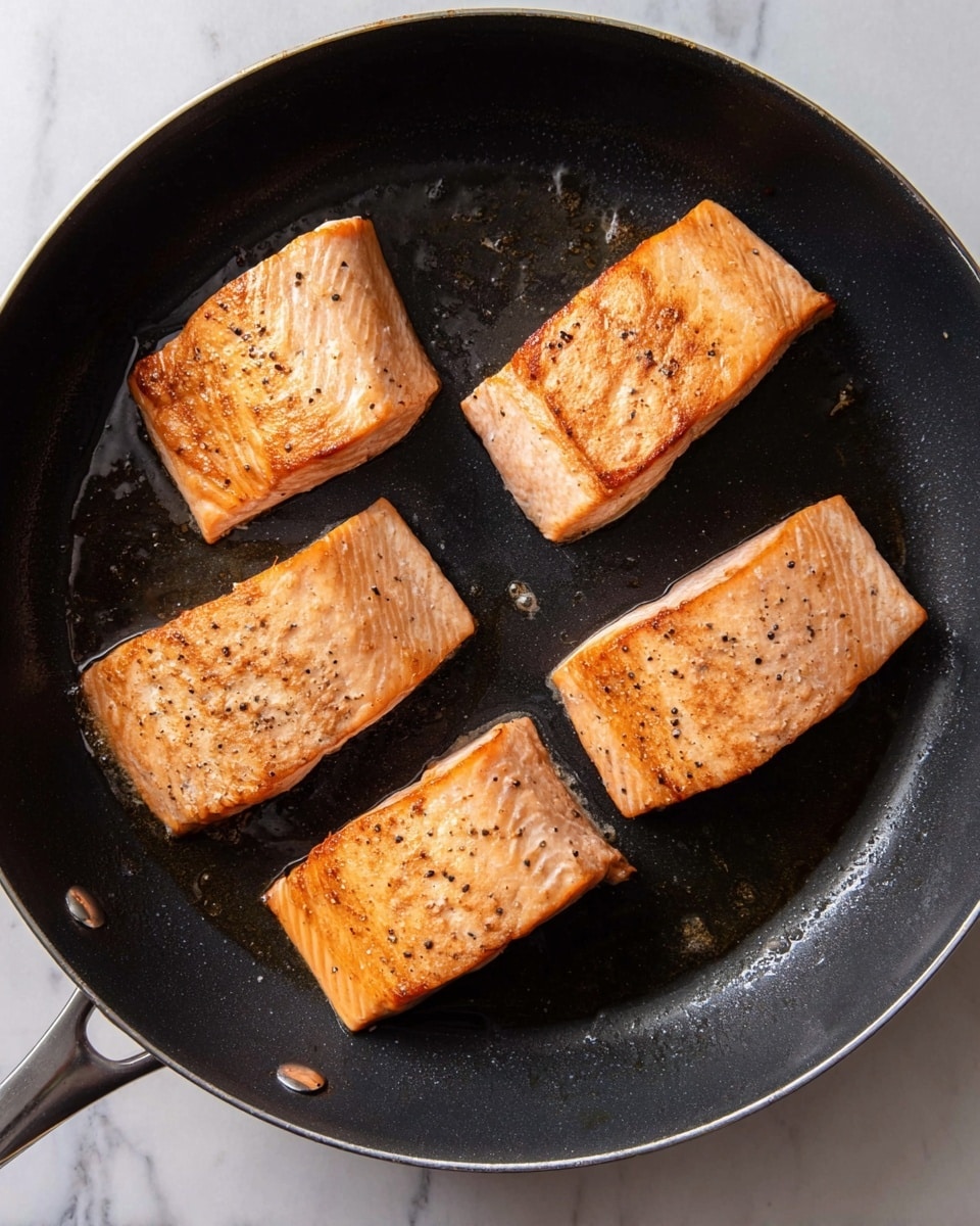 Four pieces of salmon are cooking in a black frying pan. Each piece has a golden brown, slightly crispy top with small black pepper bits scattered. The pan sits on a surface with a white marbled texture. The salmon pieces are spaced apart inside the pan, showing the shiny black pan surface with small amounts of oil around them. photo taken with an iphone --ar 4:5 --v 7