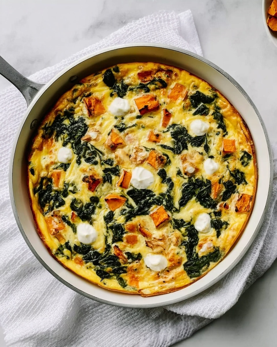 A round frittata in a white skillet with a light yellow egg base mixed with dark green spinach leaves evenly spread. Chunks of orange sweet potato are scattered on top, along with small dollops of creamy white cheese placed in a circle, adding texture. The frittata has a slightly browned edge and a soft, smooth surface. The skillet rests on a white cloth over a white marbled surface. Photo taken with an iphone --ar 4:5 --v 7