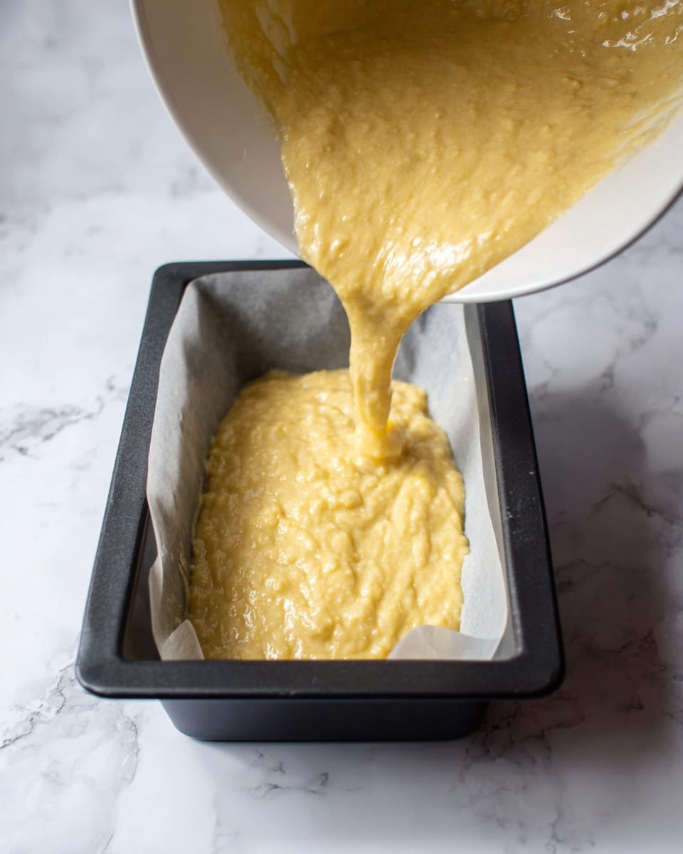 The image shows a white bowl tilted at an angle with thick, yellowish batter, which has a slightly lumpy texture, being poured into a rectangular black baking pan lined with white parchment paper. The pan is placed on a white marbled surface. The batter is flowing smoothly, covering part of the parchment paper inside the pan. photo taken with an iphone --ar 4:5 --v 7