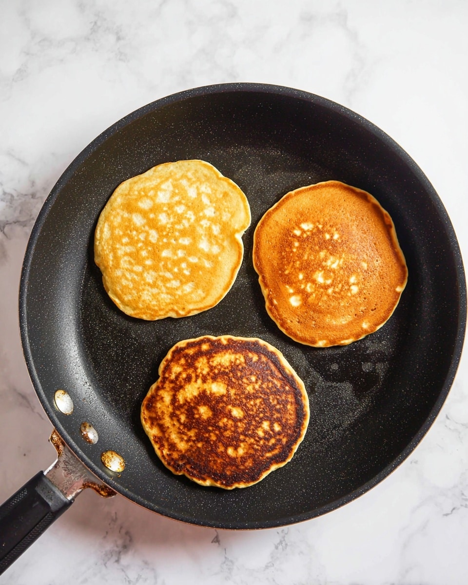 The image shows three pancakes in a black frying pan with some wear on its edges and handle. Each pancake has a golden brown top with a slightly different shade: one is lighter with some bubbles and texture, the second is evenly golden, and the third is darker with a more uneven toasted look. The frying pan sits on a white marbled surface, enhancing the contrast with the pancakes and cooking pan. Photo taken with an iphone --ar 4:5 --v 7