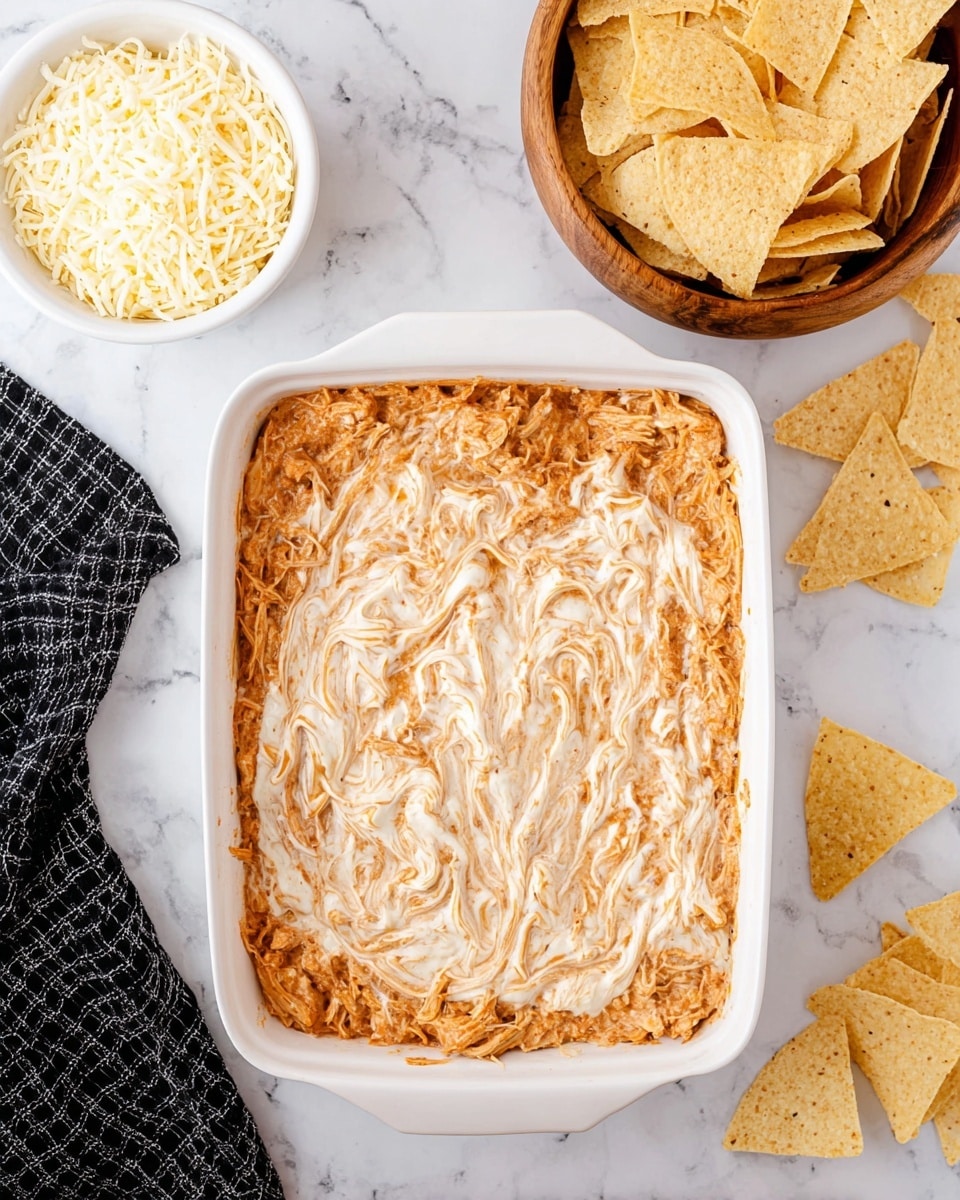 The image shows a white rectangular dish filled with a layer of shredded chicken mixed with a light reddish sauce, topped with a swirled layer of creamy white sauce spread unevenly across the surface. To the upper left of the dish, there is a white bowl filled with white shredded cheese. To the upper right sits a wooden bowl filled with light golden tortilla chips. Additional tortilla chips are placed directly on the white marbled surface near the dish. A black and white checkered cloth is partially visible at the bottom left. The photo taken with an iphone --ar 4:5 --v 7