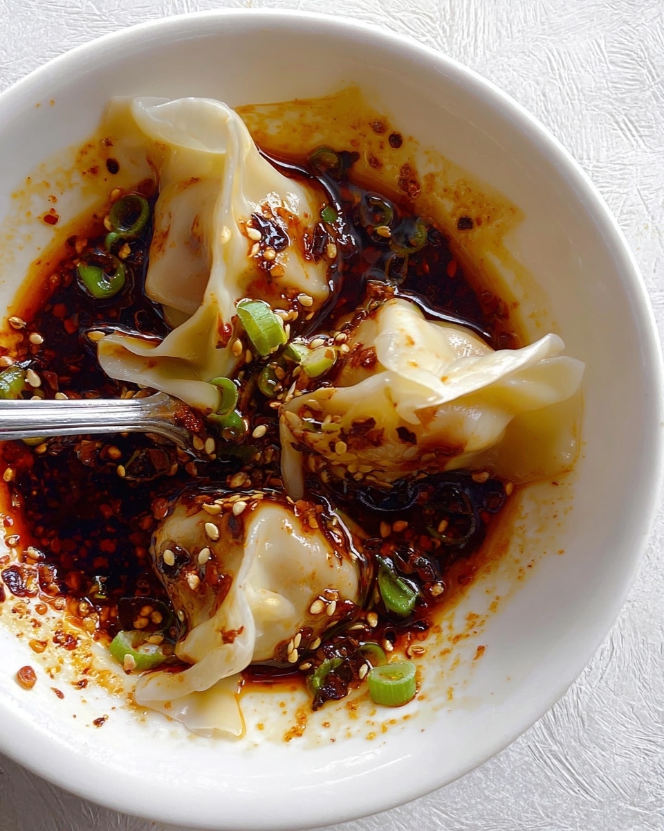 Three dumplings with smooth, pale yellow wrappers sit in a white bowl, partially folded and soaked in a dark red-brown chili oil sauce with visible bits of chili flakes and seeds. Small green onion pieces are sprinkled on top and around the dumplings. A silver fork is placed inside the bowl, piercing one dumpling. The white bowl is set on a white marbled textured surface. photo taken with an iphone --ar 4:5 --v 7