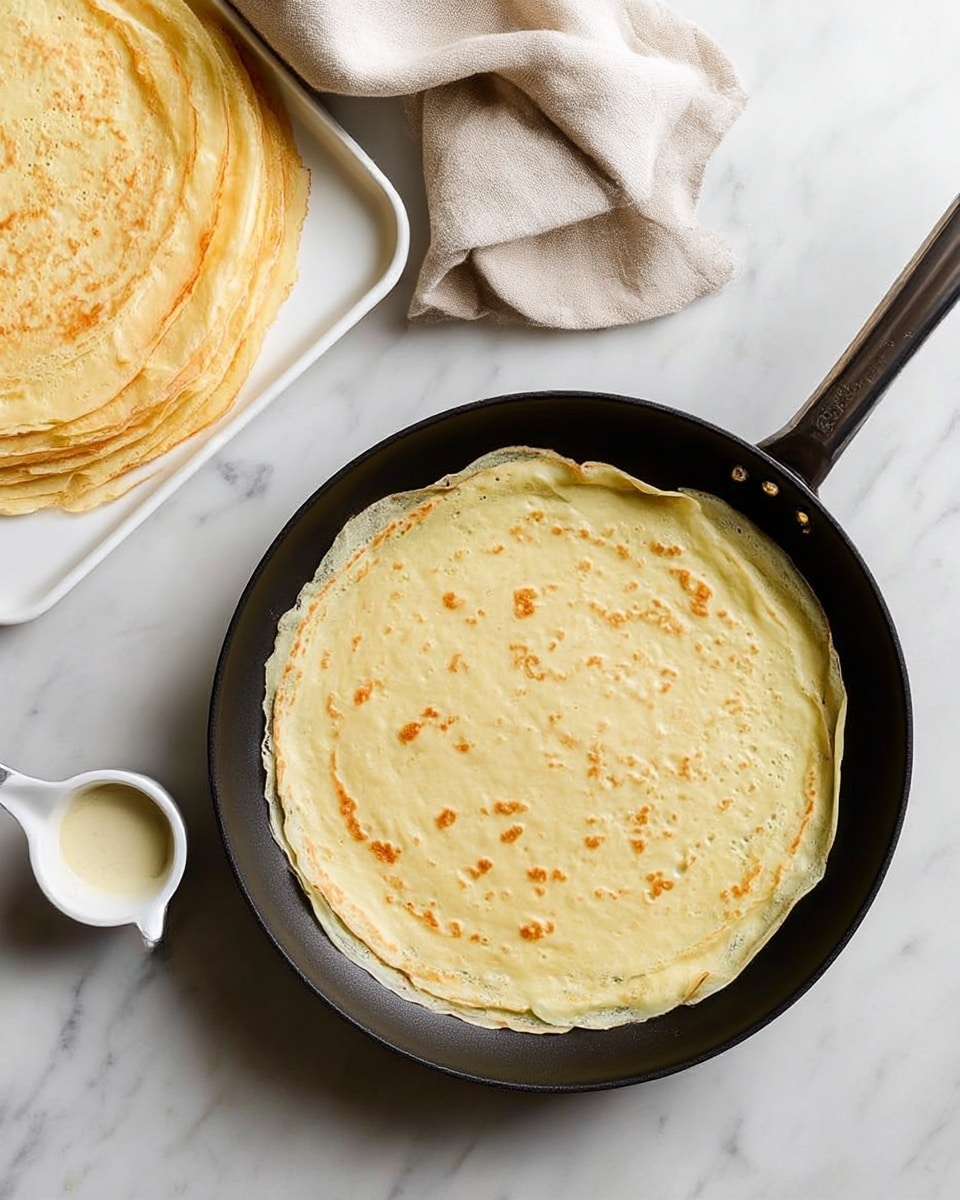 A thin, light golden crepe sits flat inside a black frying pan with a long handle, showing small browned spots and soft edges slightly curling up. To the left, there is a white tray stacked with more pale yellow crepes, each crepe's edges slightly uneven but round. A small white measuring cup with light batter inside is placed near the pan. Above the pan, a beige cloth is casually folded on a white marbled surface. The overall look is clean and soft with light colors and smooth textures. photo taken with an iphone --ar 4:5 --v 7