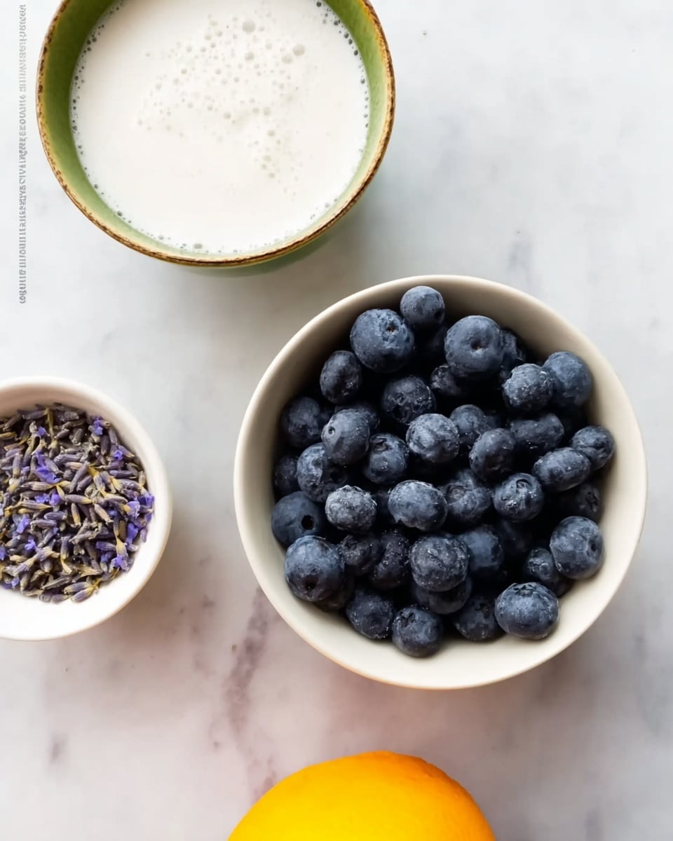 The image shows three bowls placed on a white marbled surface. The top bowl contains smooth white liquid with a slightly frothy texture, inside a green-rimmed bowl. Below it, a white bowl filled with fresh, plump blueberries, deep blue with a slightly dusty texture, dominates the middle right. To the left of the blueberries, a small white bowl holds dried lavender flowers, purple and delicate. The very bottom of the image shows the top part of an orange lemon, smooth and bright in color. photo taken with an iphone --ar 4:5 --v 7