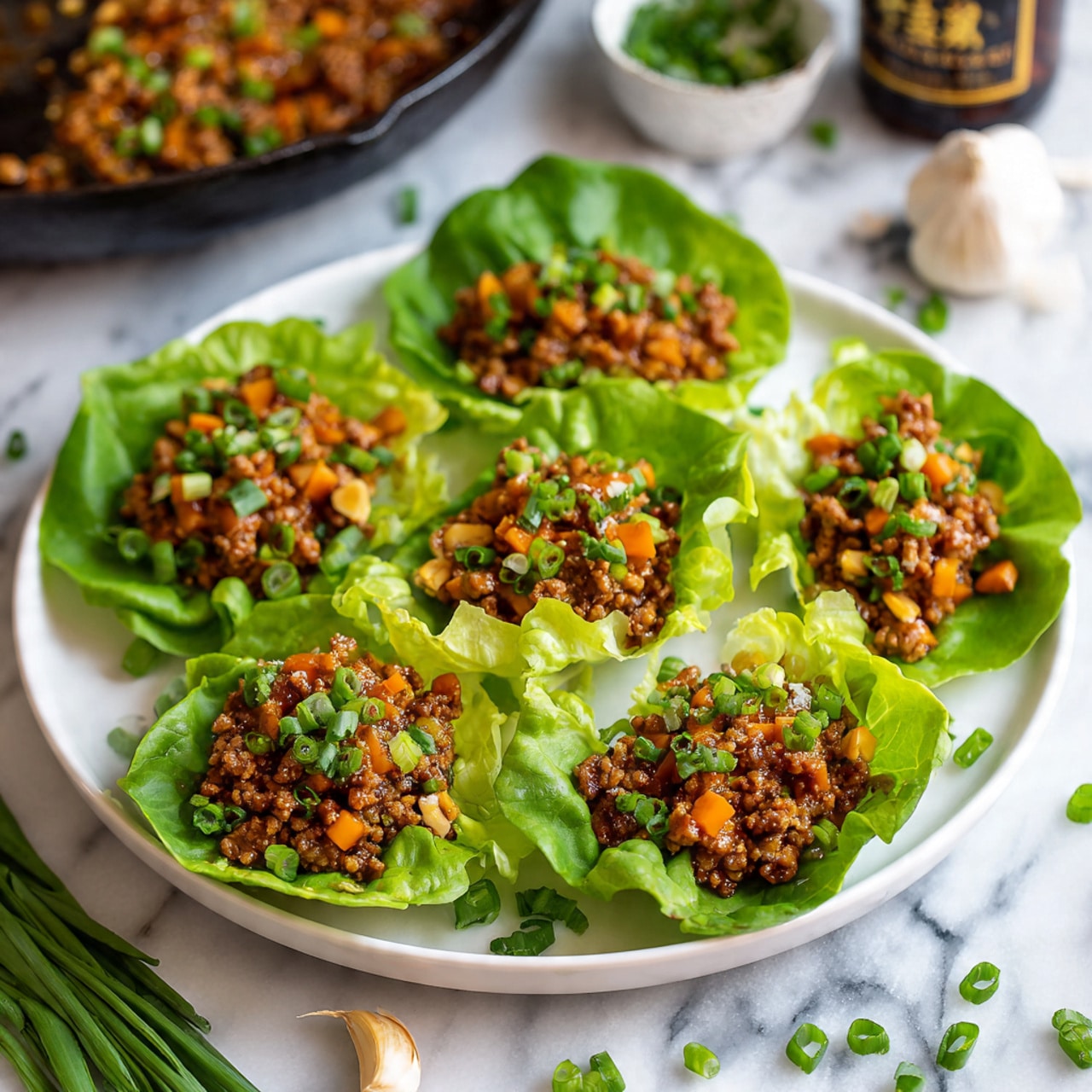 The image shows a white plate with eight green lettuce cups arranged in a circle. Each lettuce cup acts like a bowl, holding a layer of cooked minced meat mixed with small pieces of orange carrots, light-colored nuts or water chestnuts, and green chives sprinkled on top. The lettuce leaves have a bright green, slightly shiny texture with a ruffled edge. The plate is set on a white marbled surface with some scattered chopped chives and a garlic clove nearby. In the background, there is a dark pan with more of the cooked mixture and a bottle of sauce slightly out of focus. Photo taken with an iphone --ar 4:5 --v 7