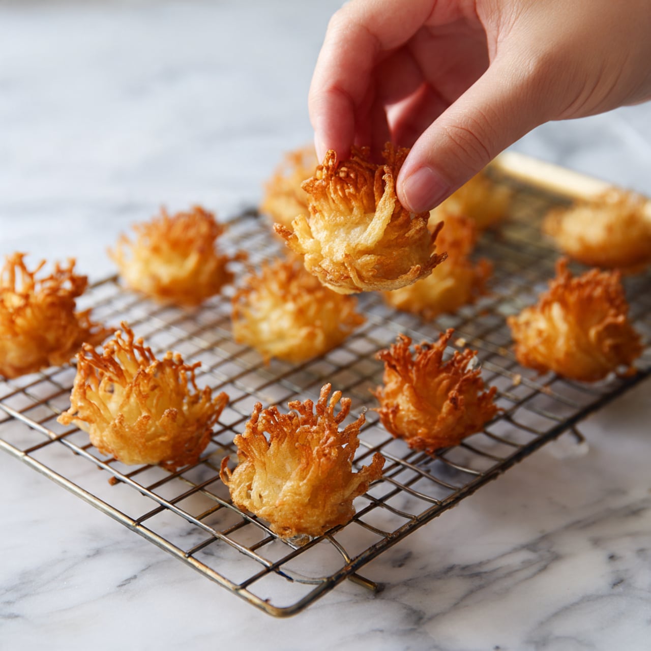 The image shows a metal cooling rack placed on a white marbled surface, holding ten pieces of deep-fried enoki mushrooms. Each fried mushroom piece has a thick, golden-brown base with multiple thin, crispy stems extending upwards, creating a crown-like shape. The mushrooms have a crunchy texture visible from their uneven and bubbled surface. A woman's hand is lifting one piece from the top left corner of the rack, showing its light and crispy detail more clearly. photo taken with an iphone --ar 4:5 --v 7