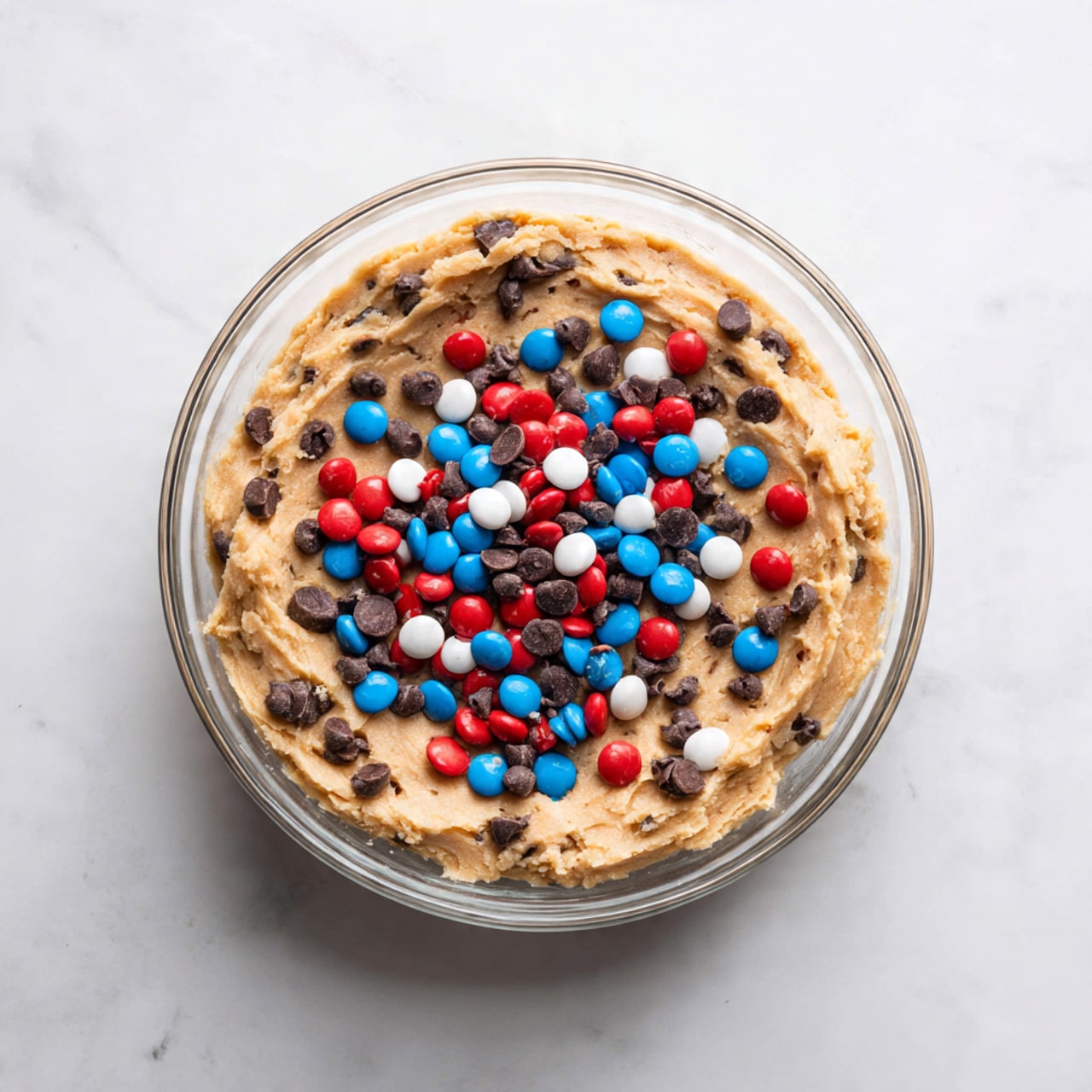 A clear glass bowl sits on a white marbled surface, filled with light brown cookie dough pressed along the sides and bottom. In the center of the bowl, there is a colorful mix of red, blue, and white candy-coated chocolates along with small dark brown chocolate chips, resting on top of the dough. The dough looks soft and textured, with the candies and chips scattered unevenly in the middle. Photo taken with an iphone --ar 4:5 --v 7