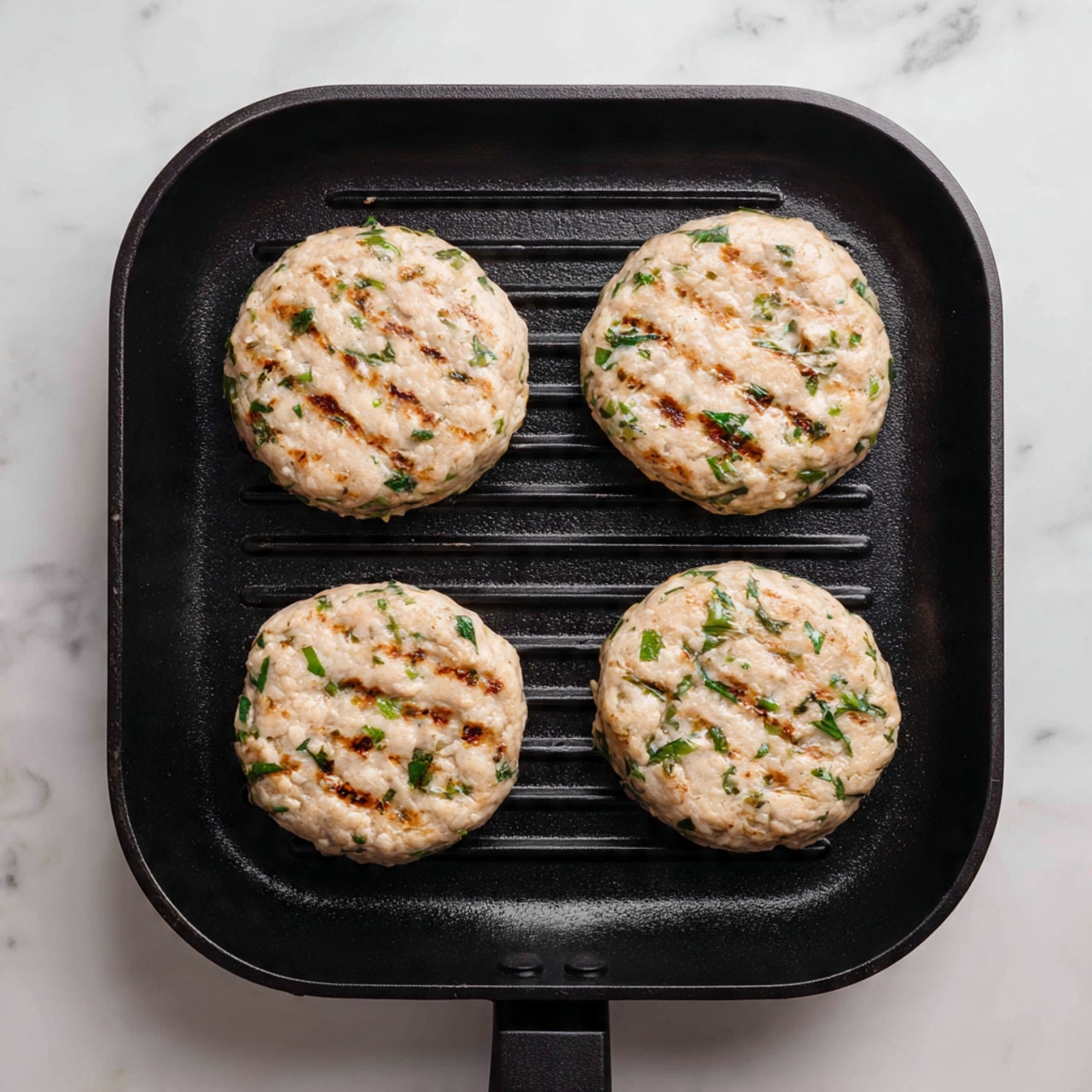 The image shows four light beige raw chicken patties with green herb bits mixed in, arranged evenly in a square shape on a black grill pan with diagonal ridges. Each patty is thick and round with uneven, rough edges and a slightly depressed center. The surface of the patties has a moist, soft texture and some pale brown spots. The grill pan has a matte finish and is placed on a white marbled surface. photo taken with an iphone --ar 4:5 --v 7
