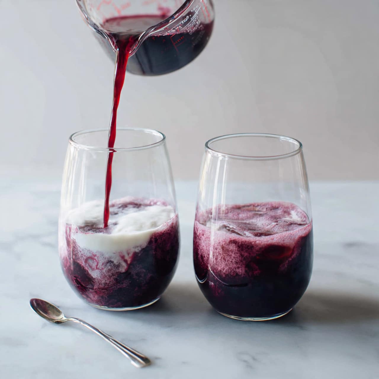 The image shows two clear glass pitchers placed side by side on a white marbled surface. Each pitcher is partially filled with a thick white liquid and a dark purple liquid is being poured into them from a Pyrex measuring cup held above. On the left, the dark purple liquid is slowly swirling down and mixing with the white liquid at the bottom, creating a marbled gradient effect. On the right, the dark purple liquid mostly fills the pitcher with a light foamy layer on top, and the white liquid is barely visible through the glass. A silver spoon lies on the white marbled surface near the bottom left of the pitchers. photo taken with an iphone --ar 4:5 --v 7