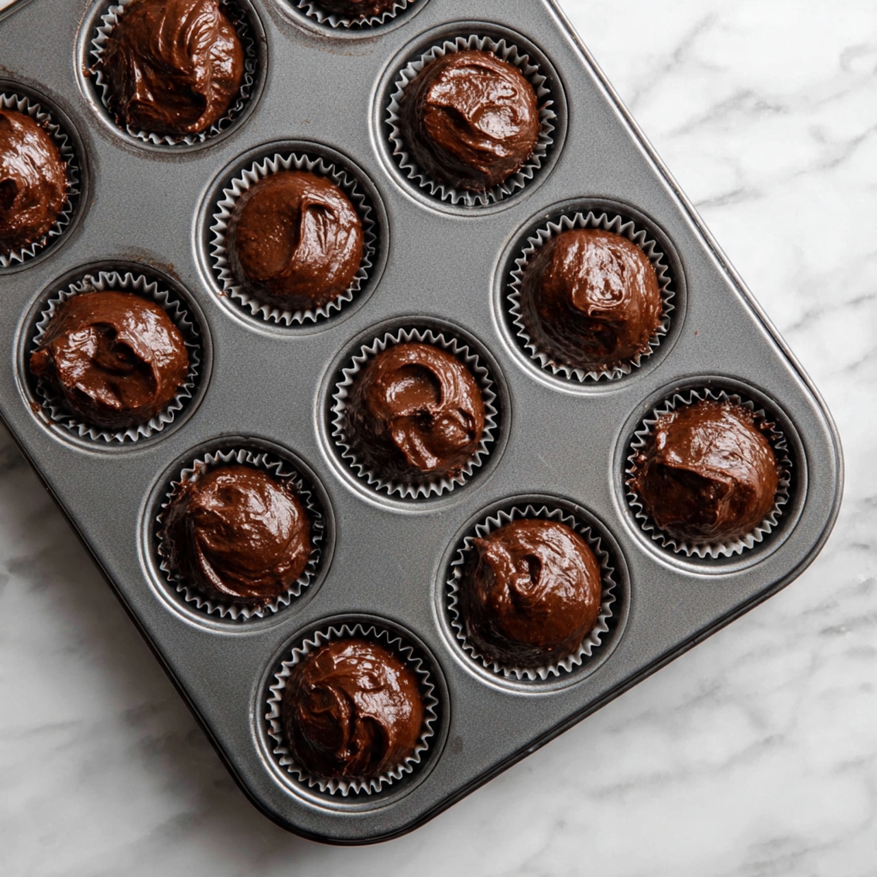 A dark gray metal mini muffin tray filled with 24 cups of thick, rich dark brown chocolate batter unevenly scooped into each cup, showing a slightly shiny and smooth surface texture. The background is a white marbled surface that contrasts with the tray's dark color, making the chocolate batter stand out. photo taken with an iphone --ar 4:5 --v 7