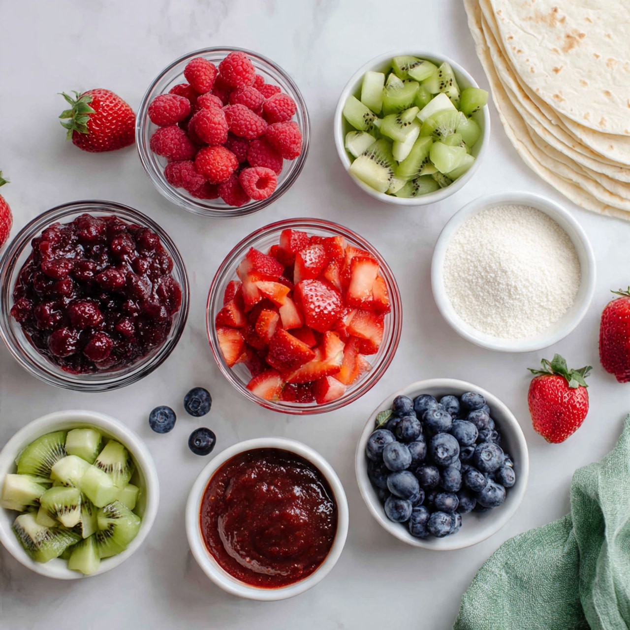 The image shows several clear and white bowls arranged on a white marbled surface, each filled with different colorful fruit and ingredients. In the center, a clear bowl holds bright red chopped strawberries, surrounded by bowls containing deep red raspberries, dark blue blueberries, green diced kiwi, light green chopped apples, and a smooth dark red fruit sauce. There is another white bowl filled with white granulated sugar nearby. A few whole strawberries and loose blueberries are scattered around the bowls, and soft white tortillas are placed in the top right corner. A green and white cloth is partially visible on the right edge. The light is natural and soft, highlighting the fresh, vibrant colors of the fruits and ingredients. photo taken with an iphone --ar 4:5 --v 7