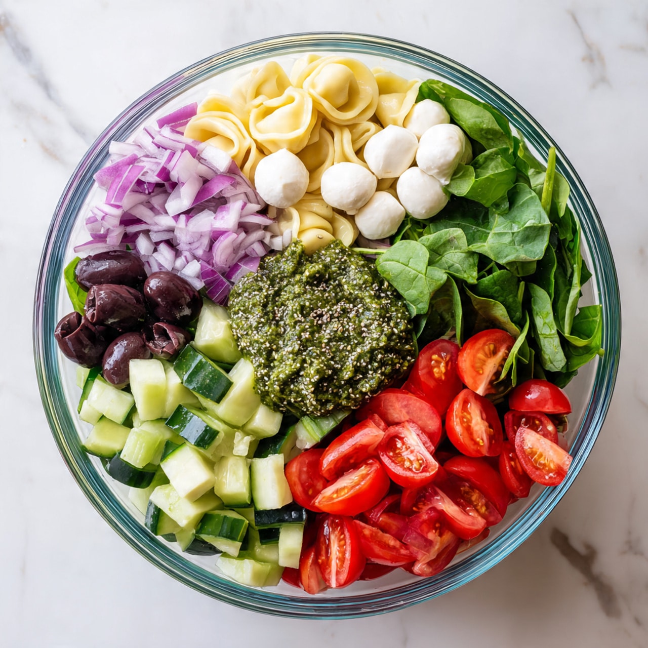 A clear glass bowl is filled with layers of fresh salad ingredients placed on a white marbled surface. Starting from the top right and moving clockwise, there is a bright green layer of leafy spinach, followed by a pile of finely chopped purple onions. Below that, a cluster of red cherry tomato halves creates a vibrant pop of color. Next, there are thick chunks of green cucumber with a slightly shiny texture. Above the cucumber, a layer of small white mozzarella balls adds a soft contrast. Beside the mozzarella, there are black sliced olives, some whole and some halved. Over the middle of the bowl is a thick, green pesto-like sauce with a chia seed garnish on top. This mix creates a colorful, fresh, and textured presentation. photo taken with an iphone --ar 4:5 --v 7