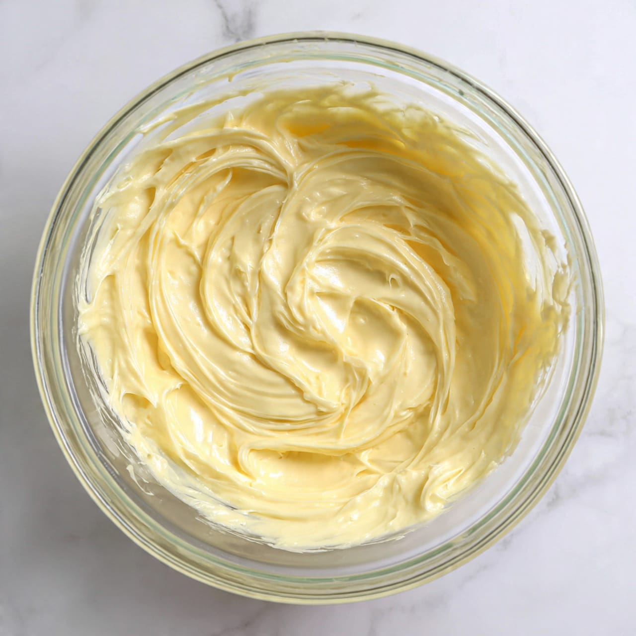 The image shows a close-up top view of one smooth creamy layer with a pale yellow color and glossy texture swirled in a clear glass bowl resting on a white marbled surface. The texture looks thick and soft with fine light reflections and slight ridges made by stirring. The edges of the bowl are barely visible, highlighting the rich mixture inside. Photo taken with an iphone --ar 4:5 --v 7