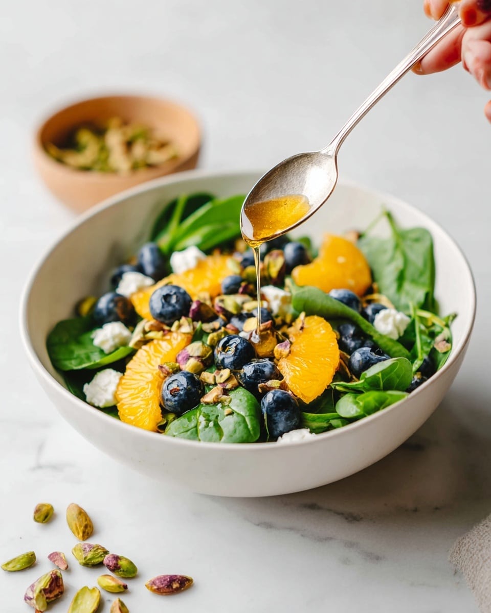 A white bowl sits on a white marbled surface filled with fresh green spinach leaves as the first layer. On top, there are bright orange segments and scattered fresh blueberries adding bursts of color. Small chunks of white cheese are spread evenly over the fruit and greens. Roasted pistachios are sprinkled all around, giving a brownish crunchy texture. A woman's hand is holding a silver spoon dripping a golden honey-like dressing over the salad. A small light brown bowl with more pistachios is partially visible in the background. Photo taken with an iphone --ar 4:5 --v 7