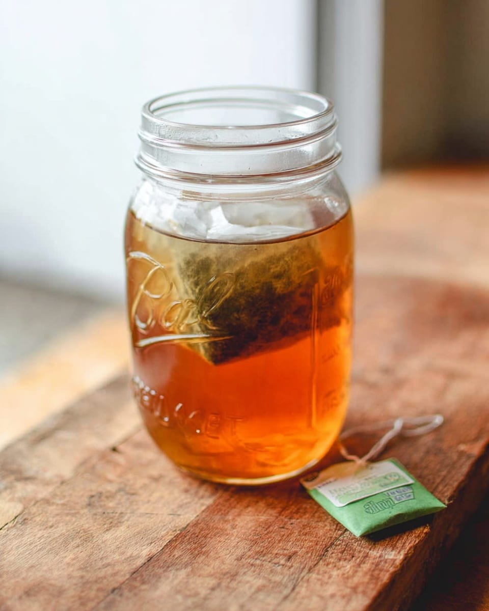 A clear glass mason jar filled with amber-colored tea sits on a wooden surface. Inside the jar, a large tea bag is partially submerged, showing a soaked and slightly darker texture at the top. Next to the jar, a green tea bag tag rests on the wooden surface. The background is softly blurred, with a clean, light tone. photo taken with an iphone --ar 4:5 --v 7